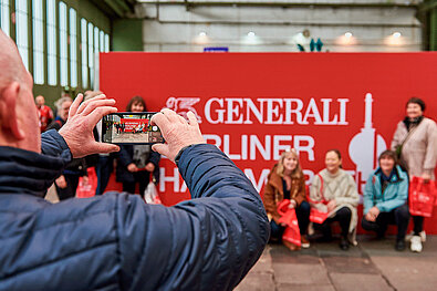 GENERALI BERLINER HALBMARATHON 2023: älterer Mann macht ein Handyfoto von einer Personengruppe vor dem Eventlogo © SCC EVENTS / Steffen Hartz