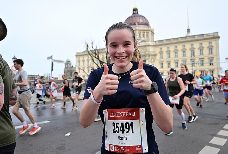 GENERALI BERLINER HALBMARATHON: Läuferin lächelt in die Kamera und zeigt zwei Daumen hoch, im Hintergrund das Berliner Schloss © SCC EVENTS / Petko Beier